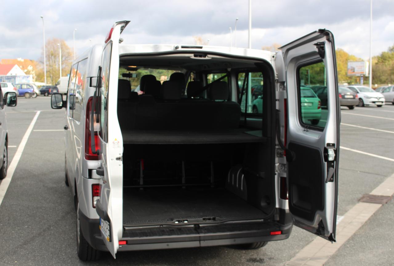 Srebrny bus z otwartymi tylnymi drzwiami, widoczna pusta przestrzeń ładunkowa z półką, zaparkowany na parkingu z innymi samochodami w tle.