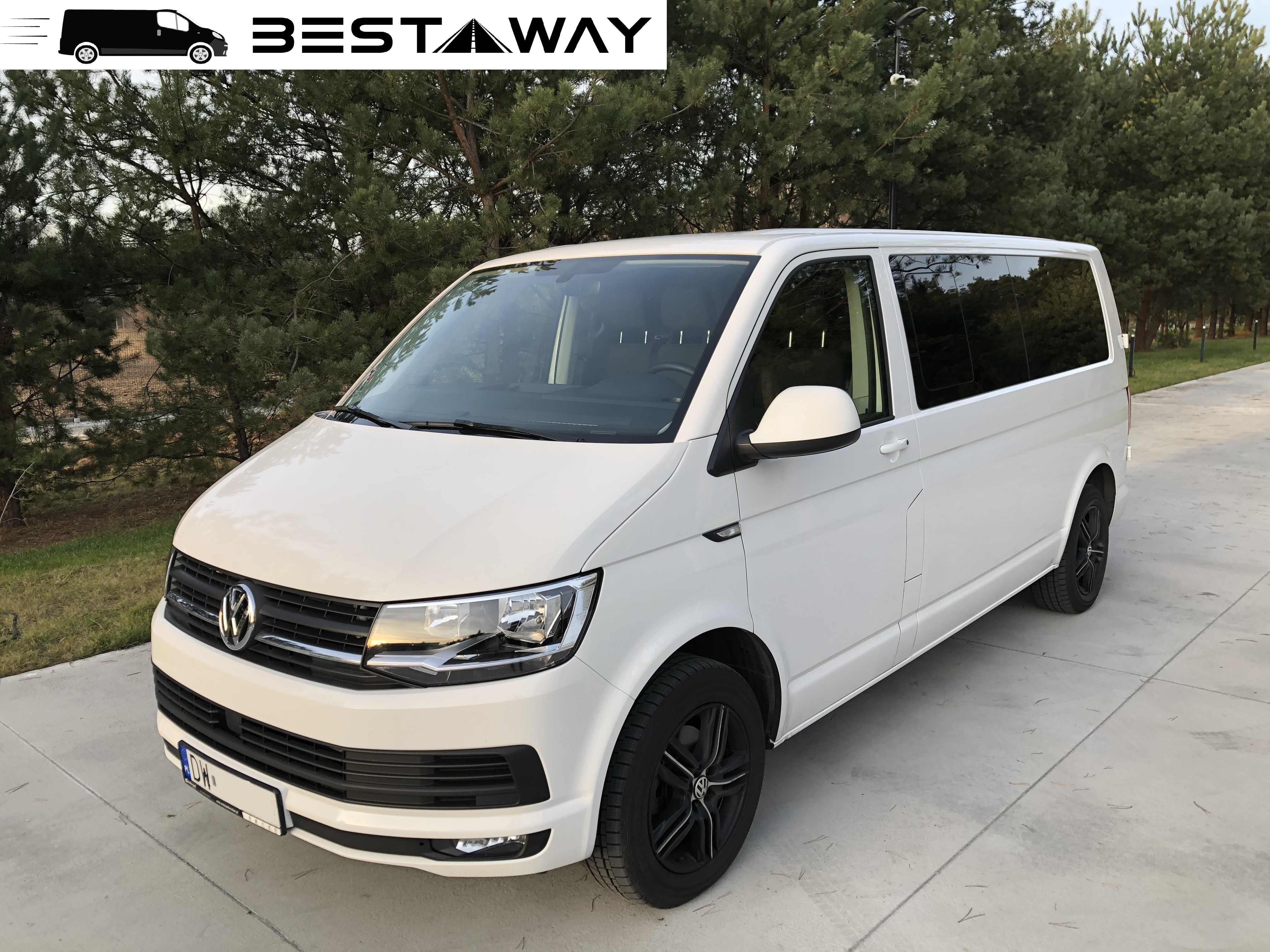 Biały bus marki Volkswagen z logo 'BESTAWAY' w tle, zaparkowany na betonowym podłożu przed zielonymi drzewami iglastymi.