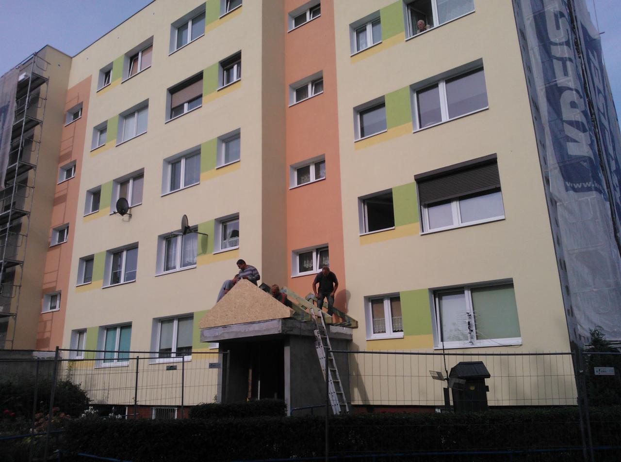 Pracownicy na dachu nad wejściem do bloku, montują drewnianą konstrukcję, elewacja bloku w odcieniach beżu, pomarańczy i zieleni, w tle rusztowania i siatka ochronna.