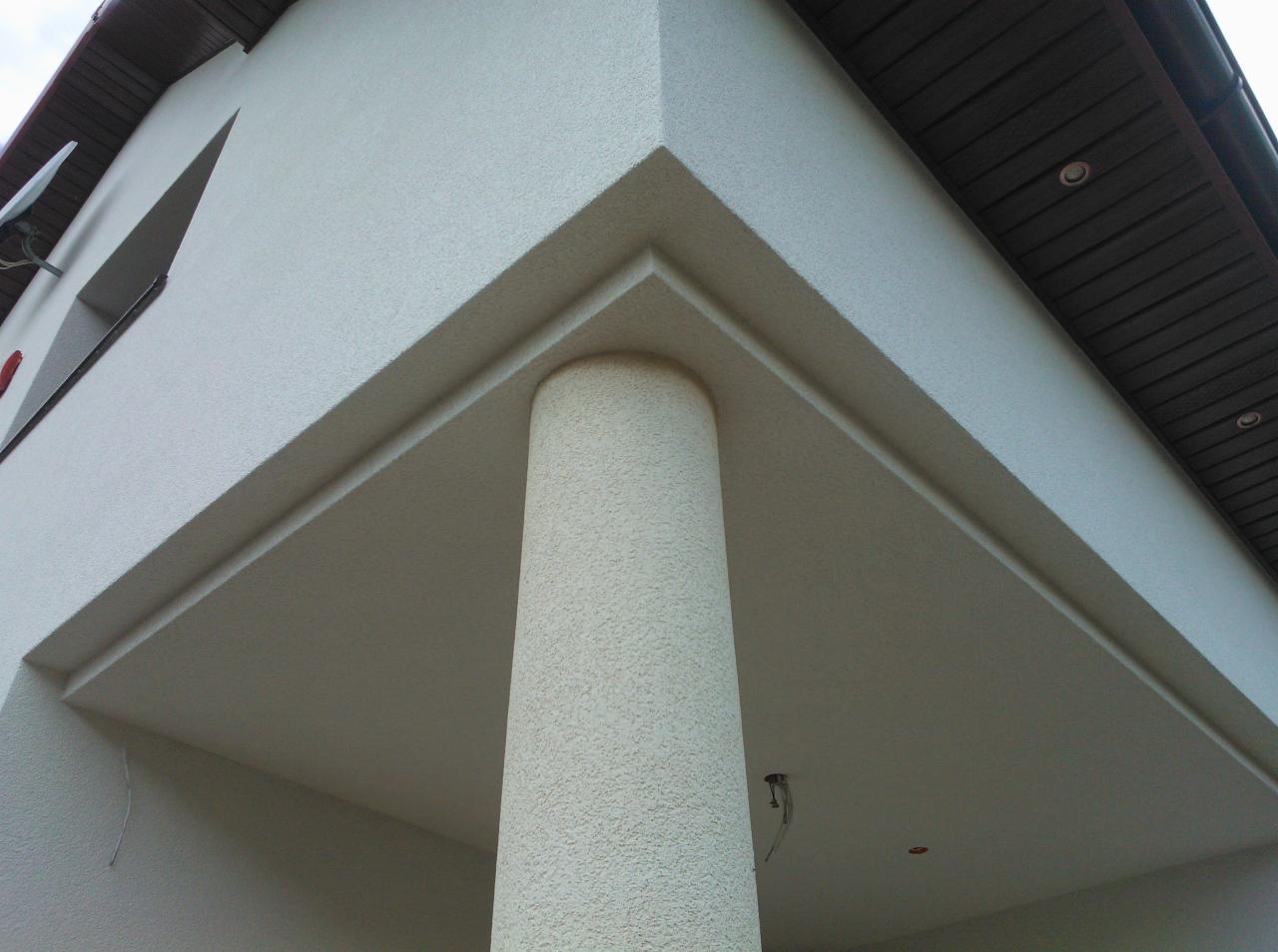 Fragment elewacji budynku z kolumną, wykończonej jasnym tynkiem strukturalnym, widok z dołu na zadaszenie i fragment dachu.