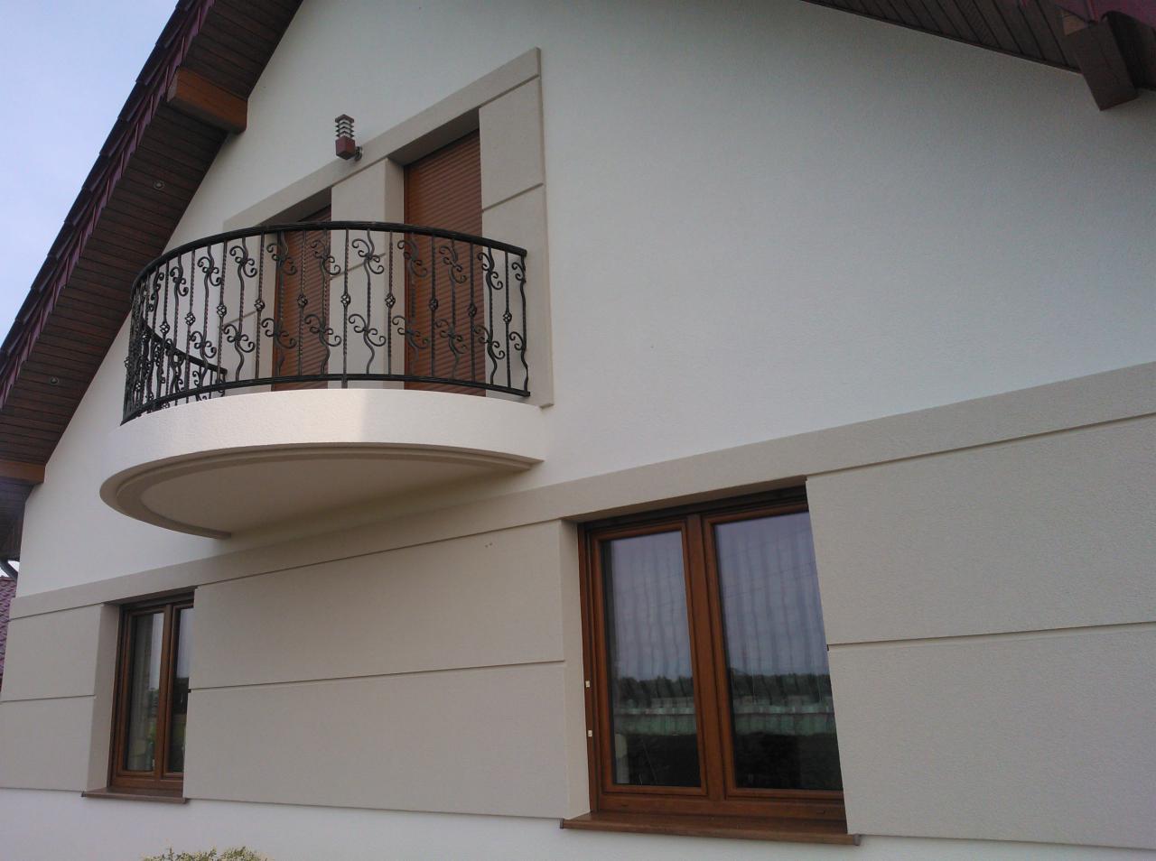 Jasna elewacja domu jednorodzinnego z balkonem o ozdobnej, metalowej balustradzie, widoczne okna z drewnianą ramą i dekoracyjne pasy na ścianie.