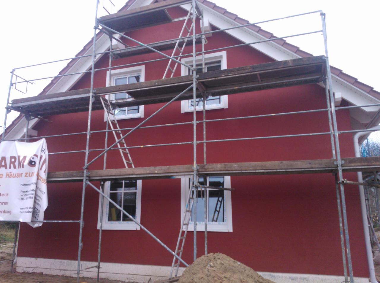 Dom jednorodzinny z czerwonymi ścianami elewacyjnymi w trakcie prac wykończeniowych, widoczne rusztowania i sterta ziemi, baner reklamowy firmy na rusztowaniu.
