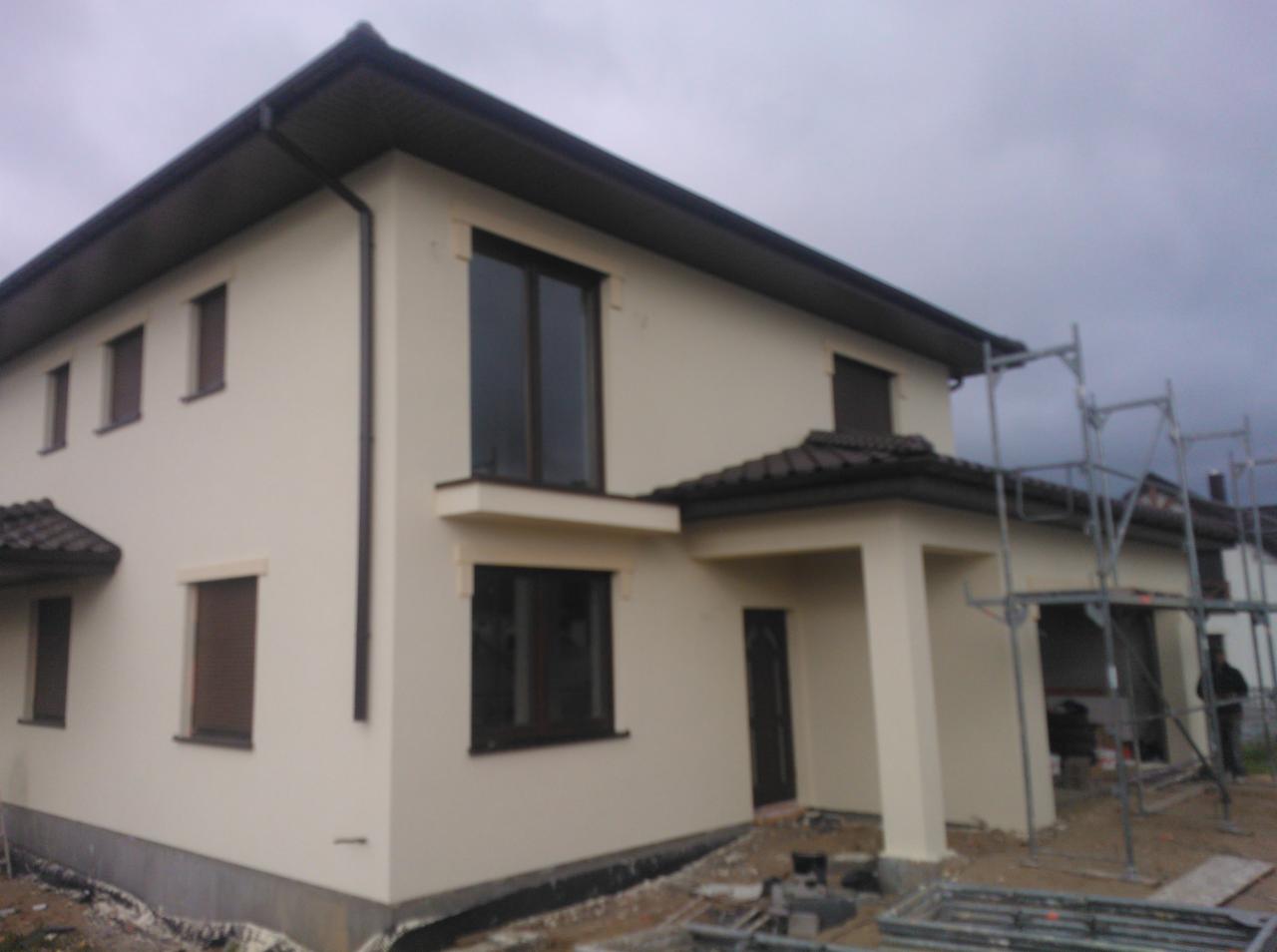 Dwukondygnacyjny dom jednorodzinny z jasną elewacją, ciemnobrązową stolarką okienną i dachem z dachówki, widoczne rusztowania przy wejściu.