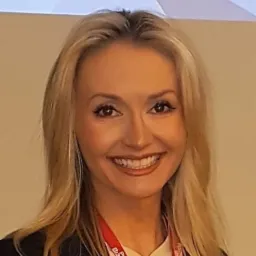 Portret uśmiechniętej kobiety z blond włosami, w ciemnej marynarce i z identyfikatorem na szyi, na jasnym tle.