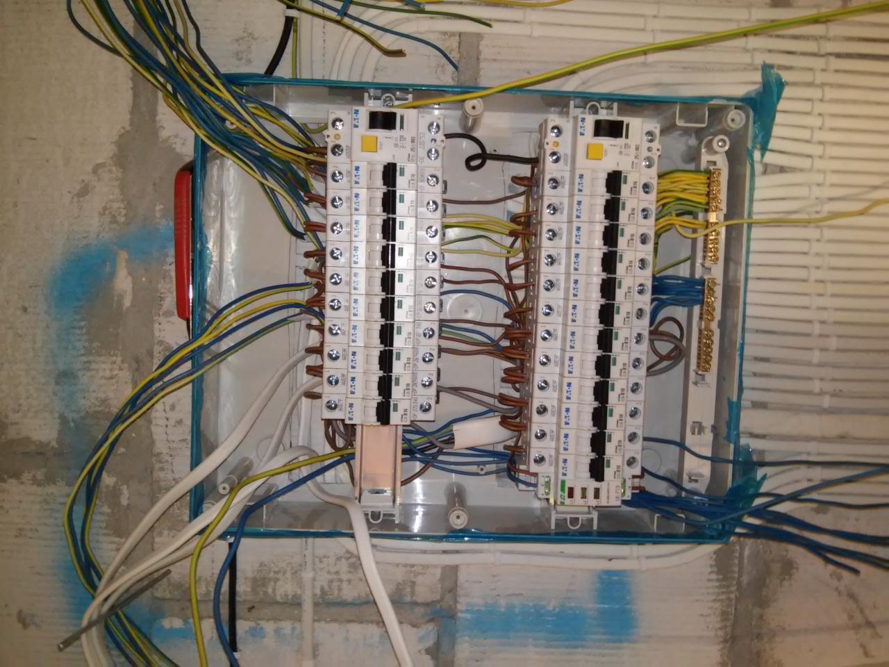 Otwarta skrzynka elektryczna z zamontowanymi bezpiecznikami Eaton, widoczne niebieskie i żółte przewody oraz ślady po malowaniu na ścianie.