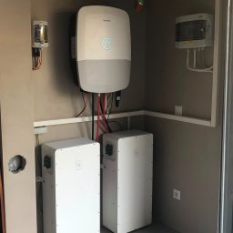 magazynenergii360 - Biały inwerter z niebieskim podświetleniem i dwa moduły magazynowania energii z logo H, zamontowane na jasnej ścianie w pomieszczeniu technicznym.
