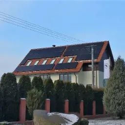 Dom jednorodzinny z panelami słonecznymi na dachu, widoczne okna dachowe i flaga na maszcie obok budynku.