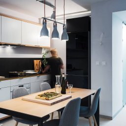 Wojciech Dziedzic FOTOGRAFIA - Nowoczesna kuchnia z jadalnią: białe szafki, czarny panel, drewniany blat, stół z pizzą i szare krzesła. Lampa sufitowa z trzema kloszami. W tle osoba przy zlewie.