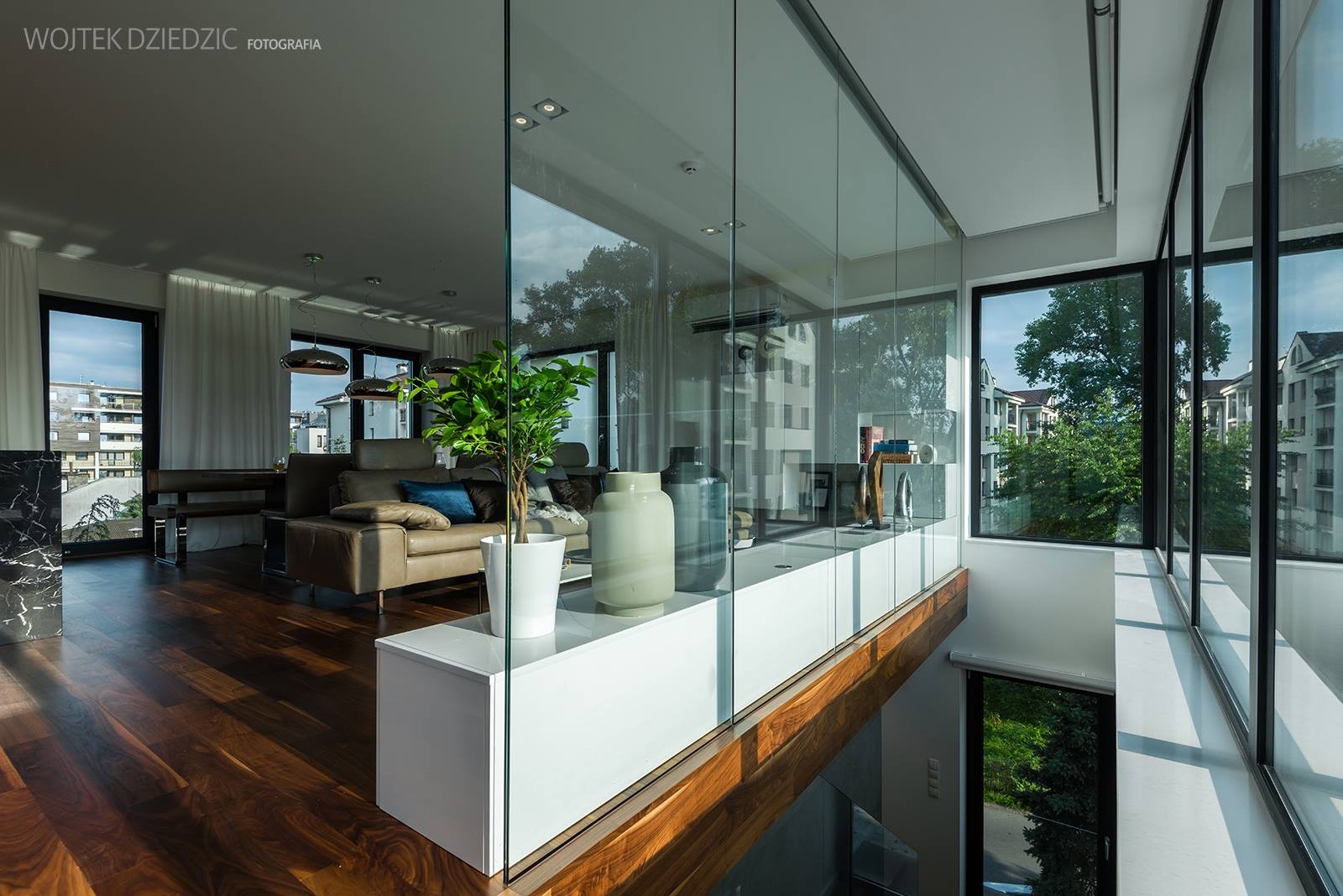 Nowoczesne wnętrze z drewnianą podłogą, skórzaną sofą i szklaną balustradą. Widok na zieleń za oknem, podkreślony przez rośliny w doniczkach. Minimalizm i elegancja.