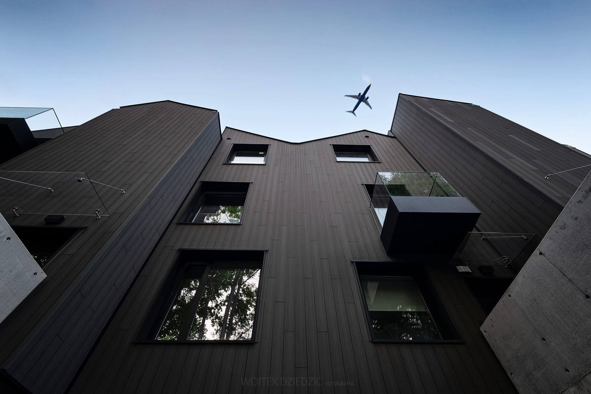 Nowoczesna, ciemna fasada budynku z drewnianym wykończeniem, widziana z dołu, z samolotem na niebie. Nietypowy kąt ujęcia.