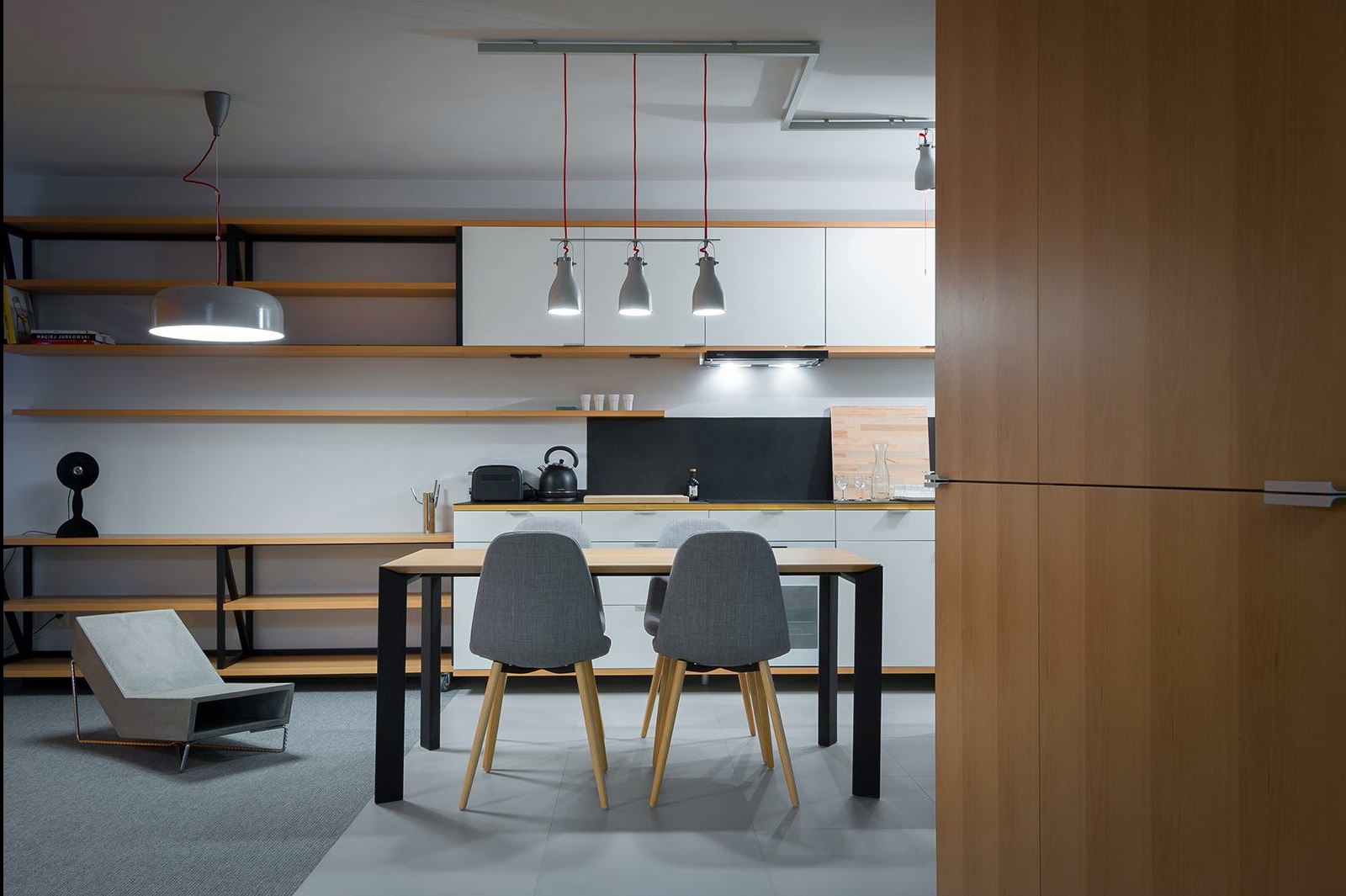 Nowoczesna kuchnia z jadalnią: minimalistyczne białe szafki, drewniane półki, szare krzesła przy stole i designerskie lampy wiszące na czerwonych kablach.