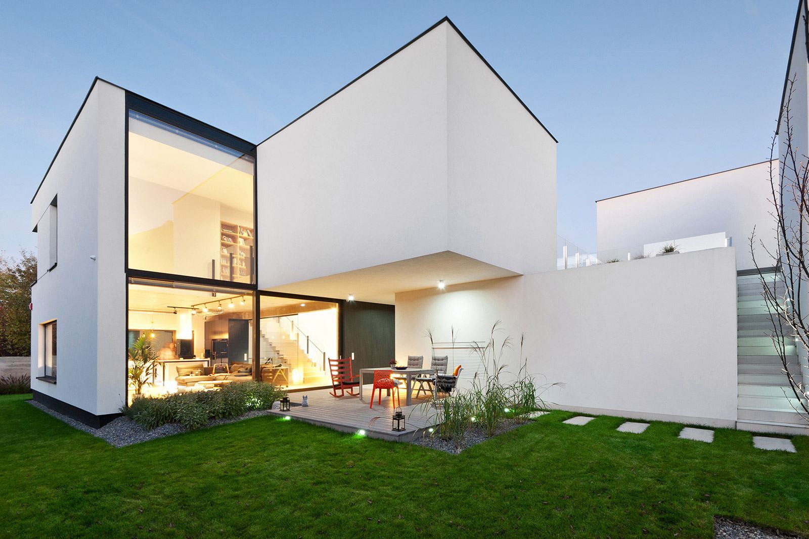 Nowoczesny dom z dużymi przeszkleniami i tarasem. Widok na wnętrze z białymi schodami i salonem. Oświetlony ogród z meblami.