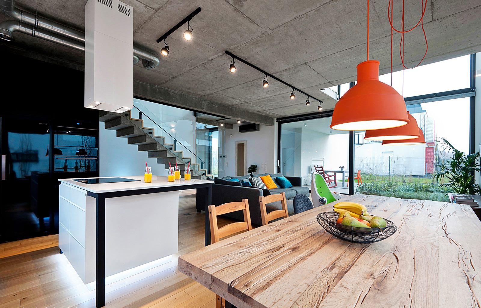 Nowoczesne wnętrze domu z betonowym sufitem, drewnianym stołem i pomarańczowymi lampami. Widok na ogród przez duże okno.