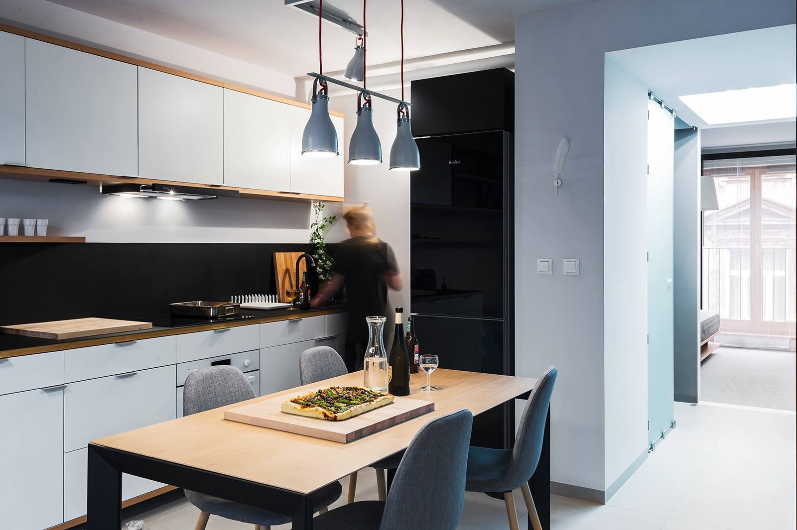 Nowoczesna kuchnia z jadalnią: białe szafki, czarny panel, drewniany blat, stół z pizzą i szare krzesła. Lampa sufitowa z trzema kloszami. W tle osoba przy zlewie.