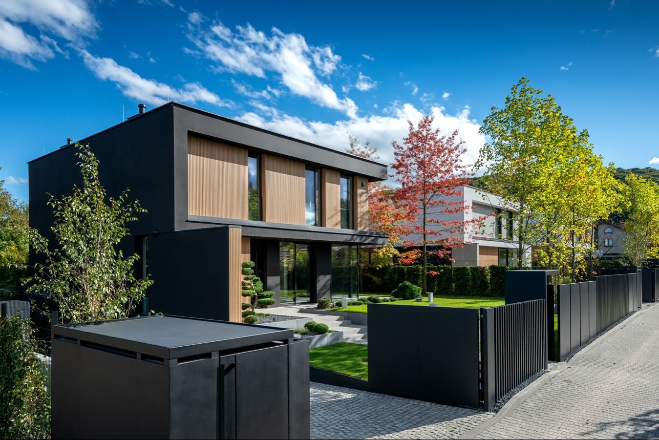 Nowoczesny dom z elementami drewna, czarną elewacją i zadbanym ogrodem. Błękitne niebo z obłokami w tle. Widok z poziomu ulicy, brama wjazdowa na pierwszym planie.