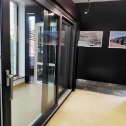 Otwarta, przesuwna, antracytowa rama okienna z widocznym uchwytem, na tle ciemnej ściany z oświetleniem punktowym i plakatami.