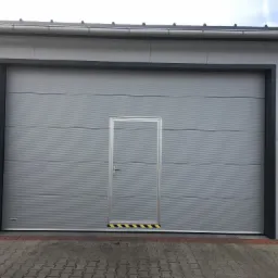 Szara brama garażowa segmentowa z wbudowanymi drzwiami wejściowymi, widoczne czarno-żółte pasy ostrzegawcze na progu, fasada budynku z metalowych paneli, kostka brukowa na pierwszym planie.