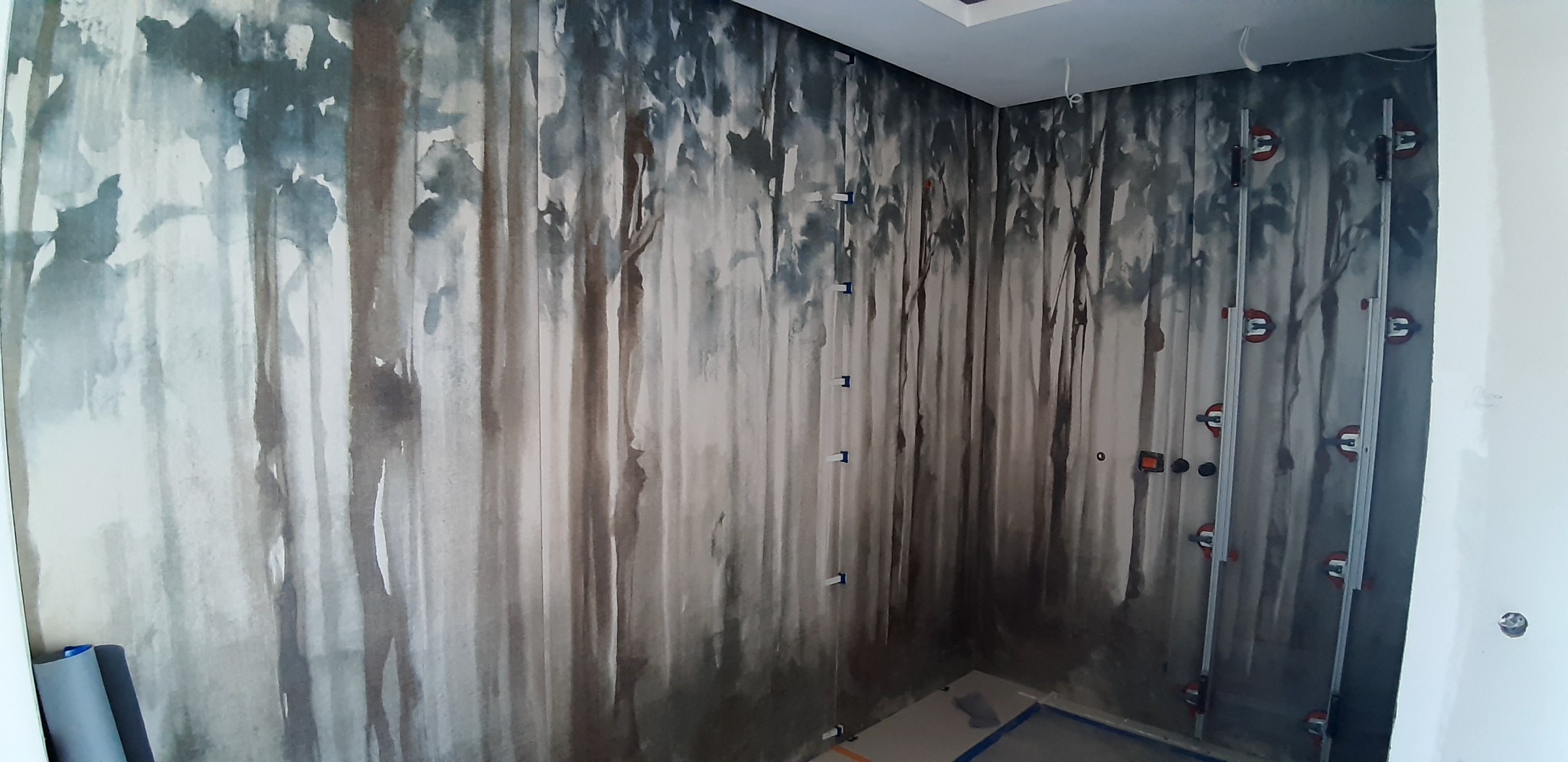 Wnętrze pokoju z tapetą imitującą las w akwarelowym stylu, widoczne narzędzia glazurnicze i listwy do mocowania tapety, podłoga częściowo zabezpieczona kartonem