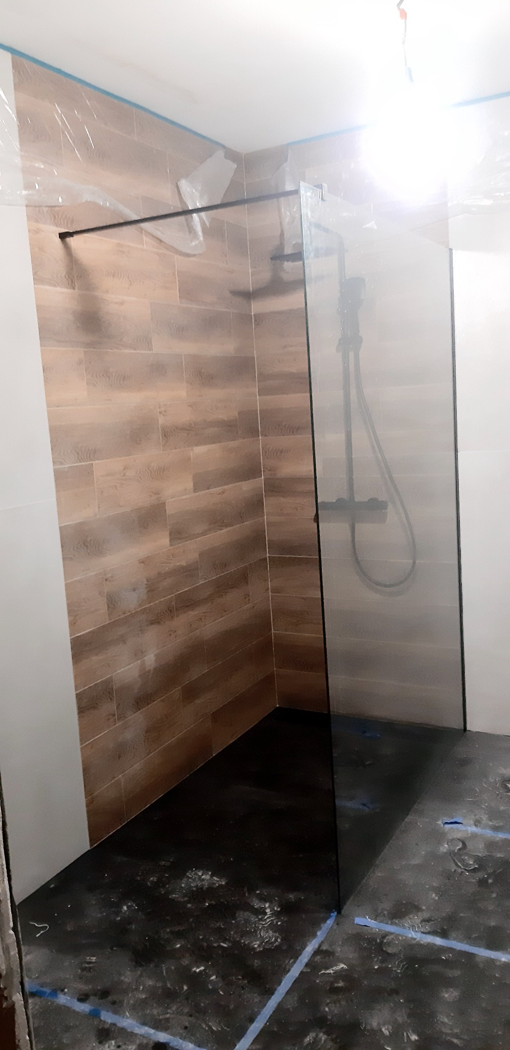 Nowoczesna kabina prysznicowa typu walk-in z czarną armaturą i panelami imitującymi drewno, w trakcie montażu, zabezpieczona folią malarską.