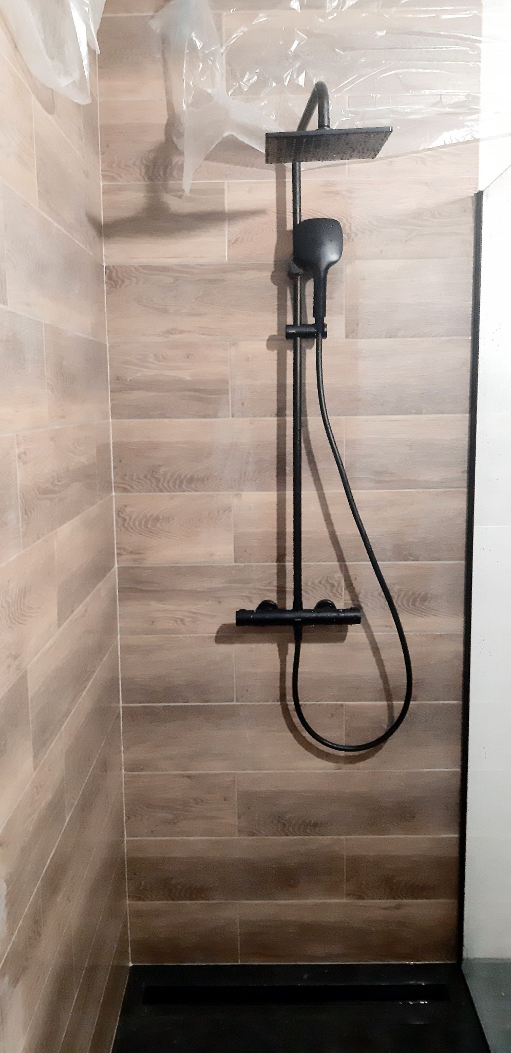 Wykończona kabina prysznicowa z czarną armaturą i drewnopodobnymi płytkami na ścianach, zabezpieczona folią malarską.