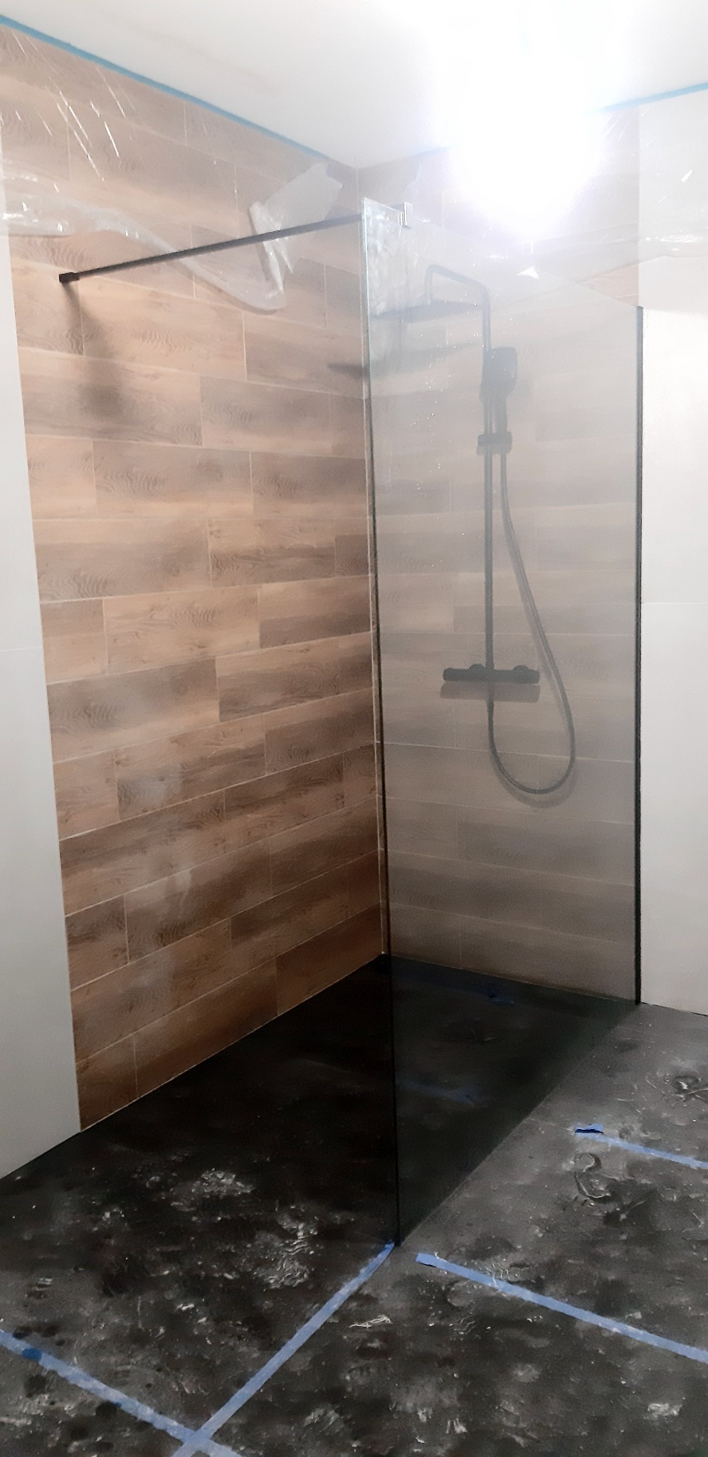 Nowoczesna kabina prysznicowa typu walk-in z czarną armaturą i panelami ściennymi imitującymi drewno, widoczne ślady prac wykończeniowych na podłodze.