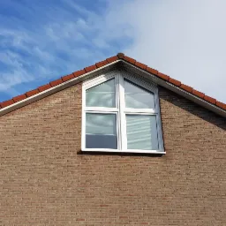 Trójkątne okno z białymi ramami w szczycie ceglanego domu pod błękitnym niebem z chmurami.