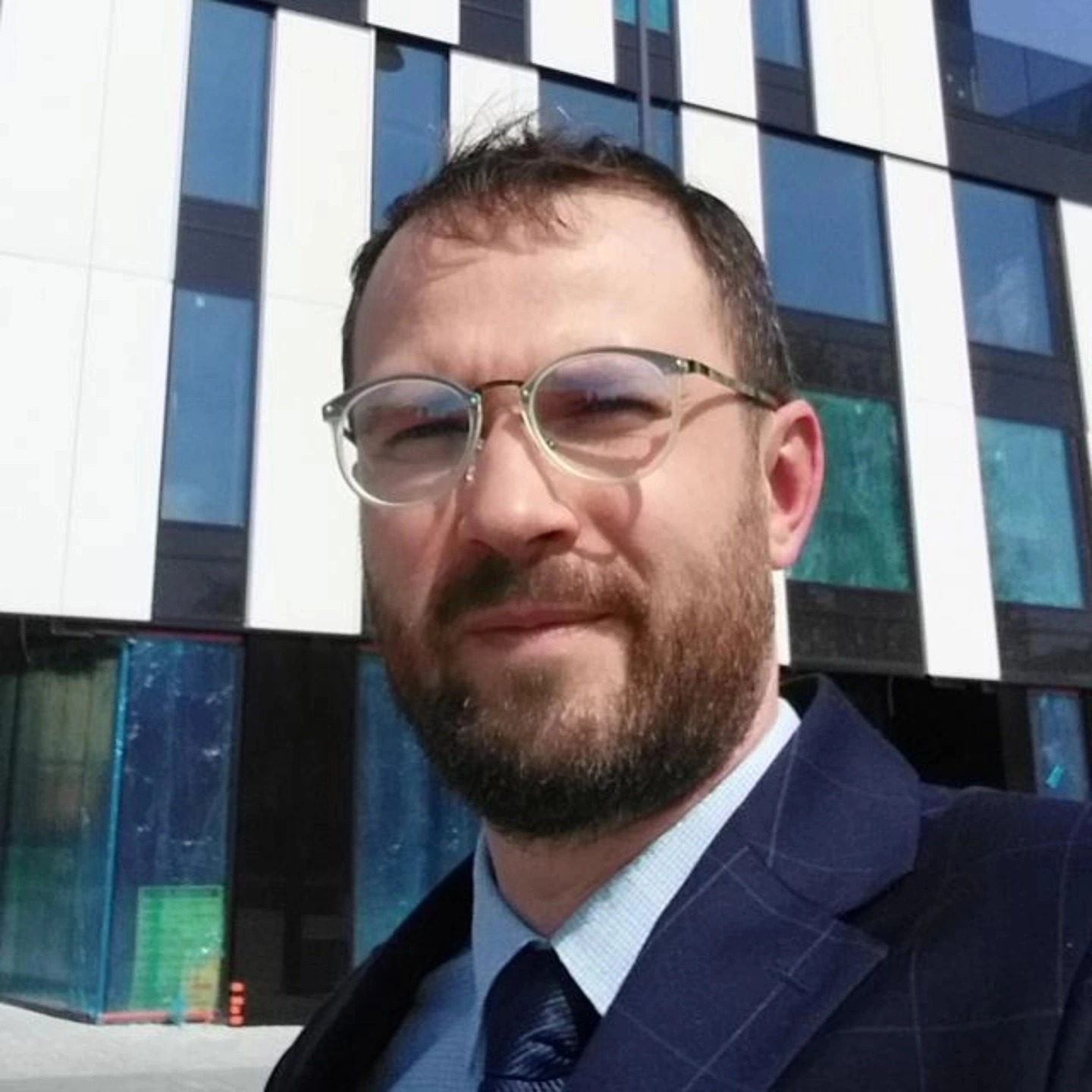 Portret mężczyzny w okularach i garniturze na tle nowoczesnego budynku z biało-czarną fasadą. Widoczny fragment niebieskiego nieba.