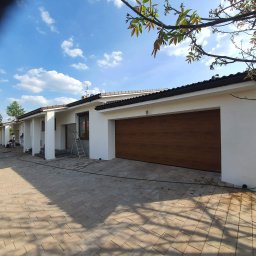 Marek Wiśniewski - Biała elewacja domu jednorodzinnego z brązową bramą garażową i kostką brukową na podjeździe, widoczne narzędzia i pracownicy podczas czyszczenia elewacji.