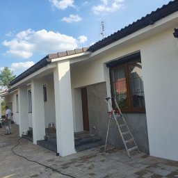 Marek Wiśniewski - Dwóch mężczyzn maluje elewację domu z drabiny, widoczne narzędzia i sprzęt do malowania, błękitne niebo z chmurami w tle.