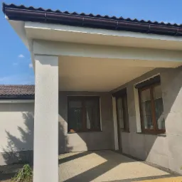 Zadaszone wejście do domu z elewacją wykończoną szarymi płytami, brązowe okna i dachówka, jasna kolumna i podłoga z kostki.