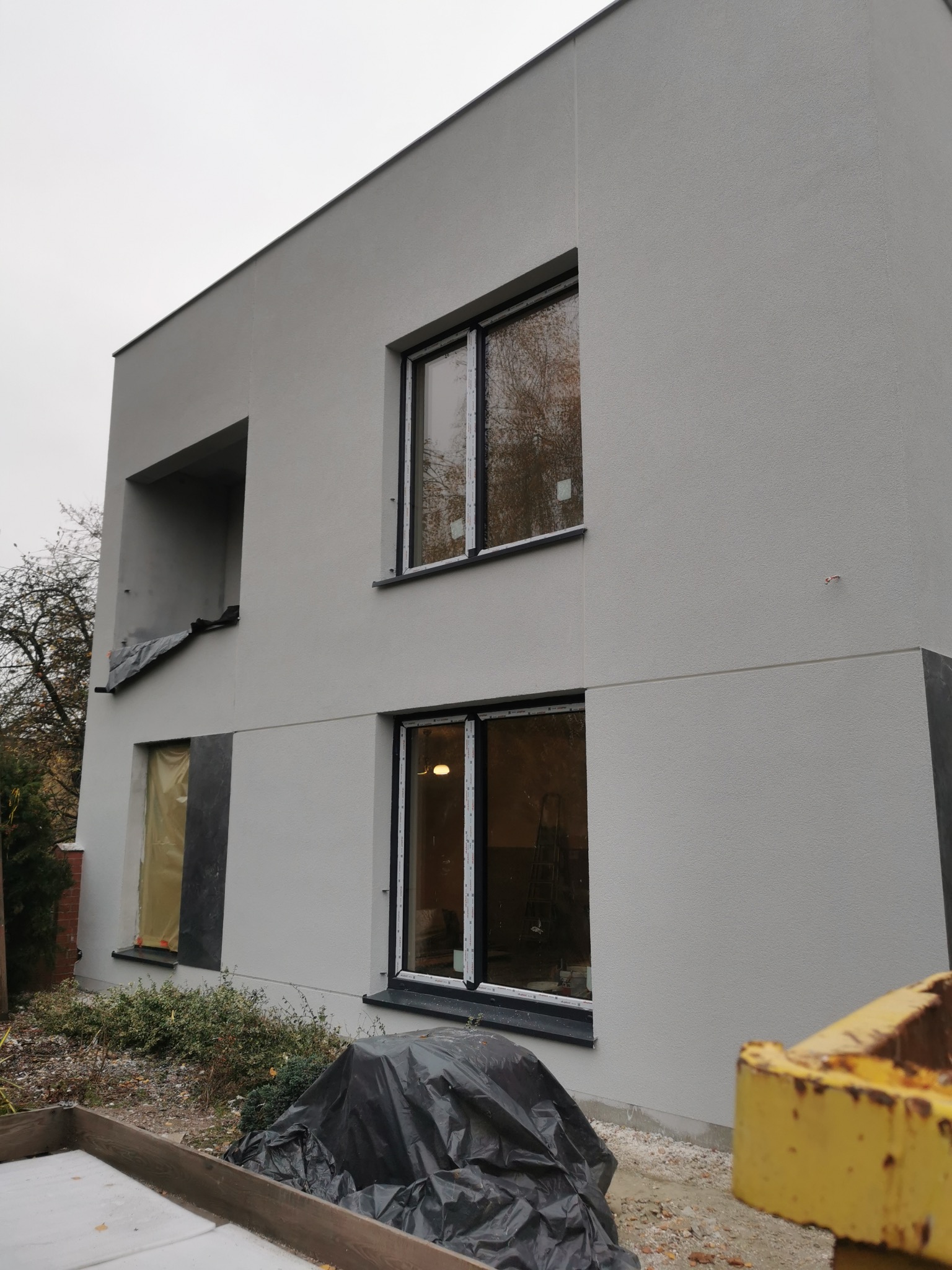 Dwupiętrowy budynek z szarą elewacją po tynkowaniu, z oknami w czarnych ramach; jedno okno zabezpieczone folią ochronną, widoczne ślady prac budowlanych wokół budynku.