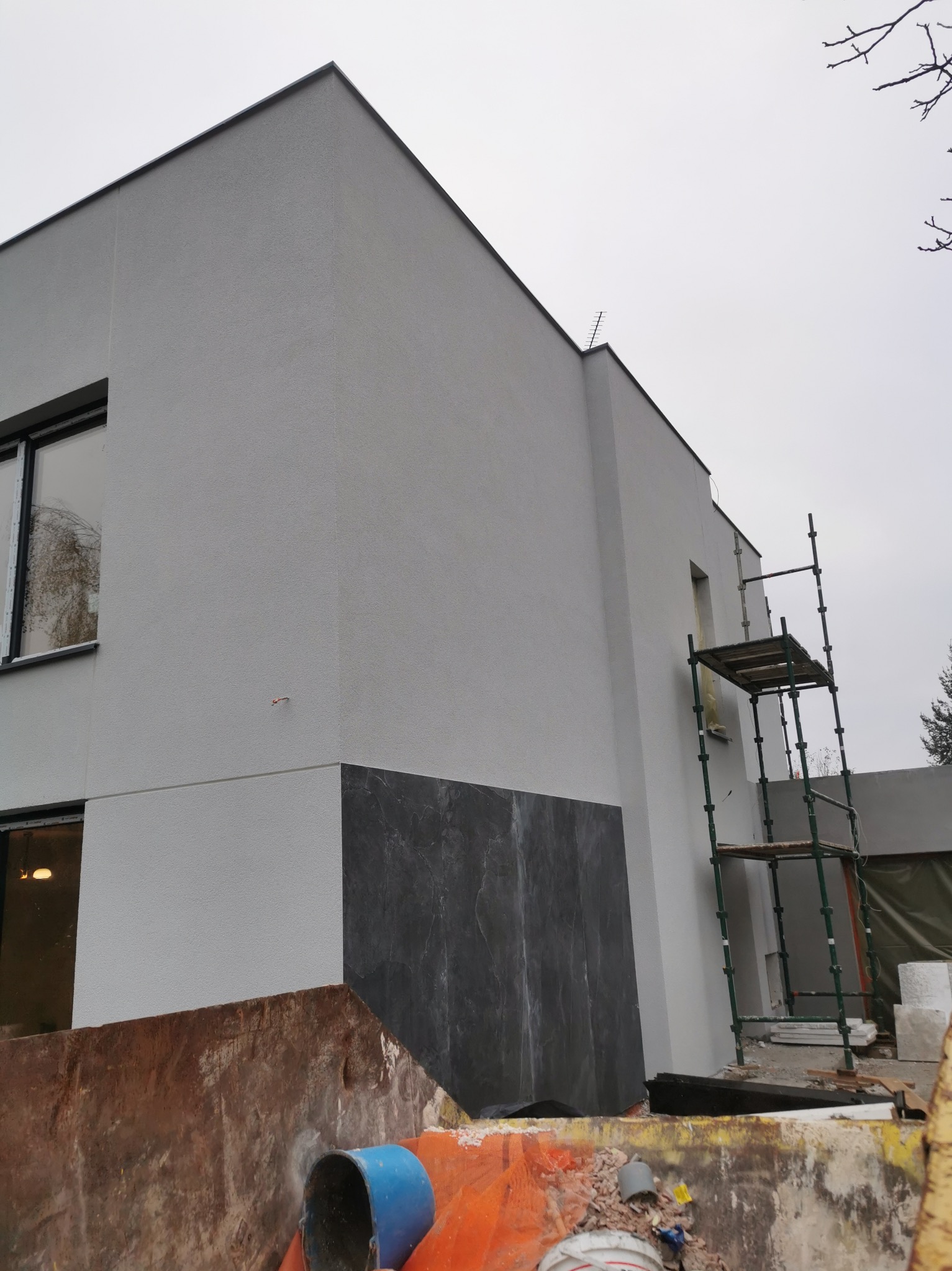 Szara elewacja budynku z fragmentem wykończonym czarnym kamieniem, widoczne rusztowanie i kontener z gruzem.