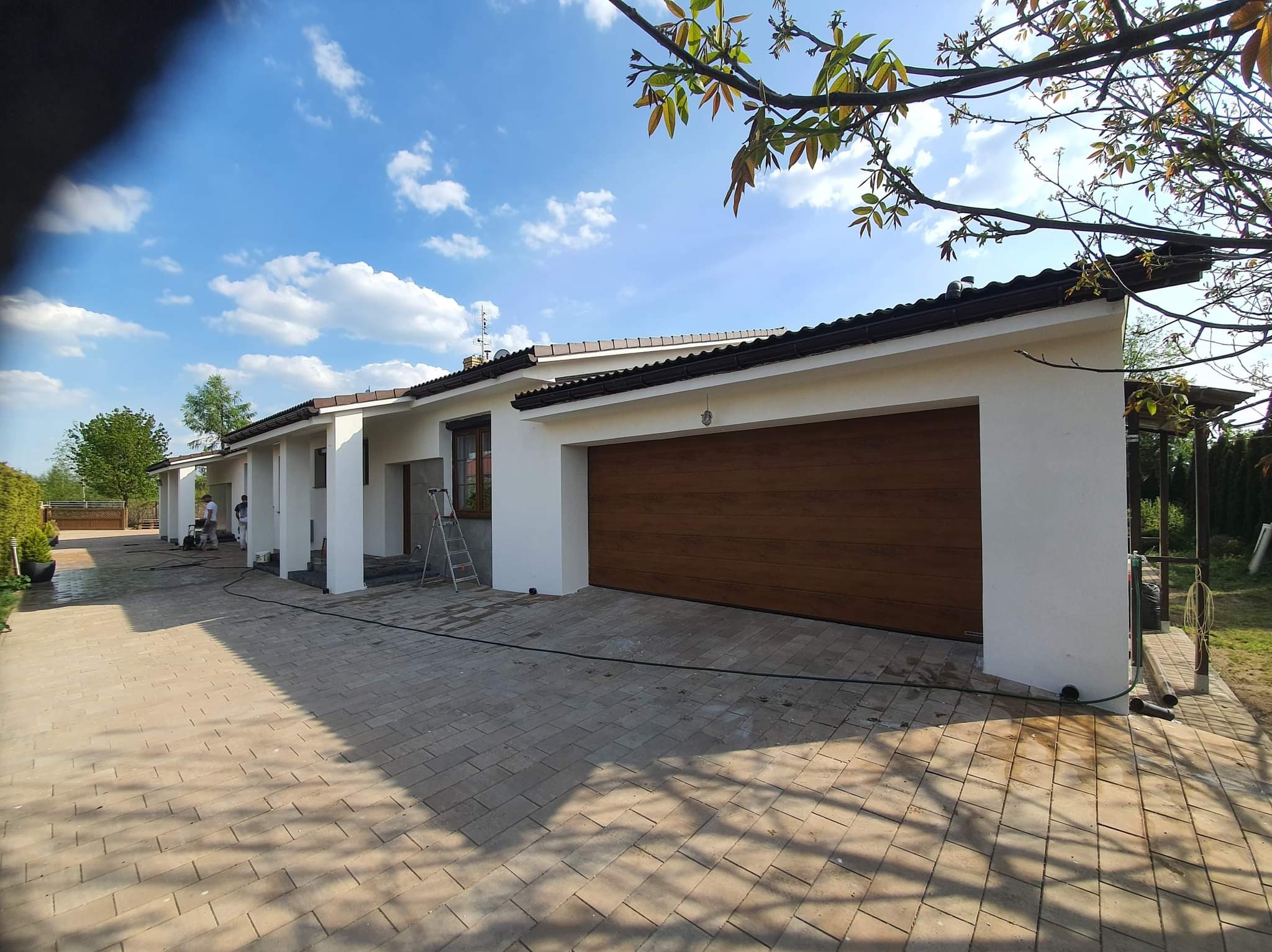 Biała elewacja domu jednorodzinnego z brązową bramą garażową i kostką brukową na podjeździe, widoczne narzędzia i pracownicy podczas czyszczenia elewacji.