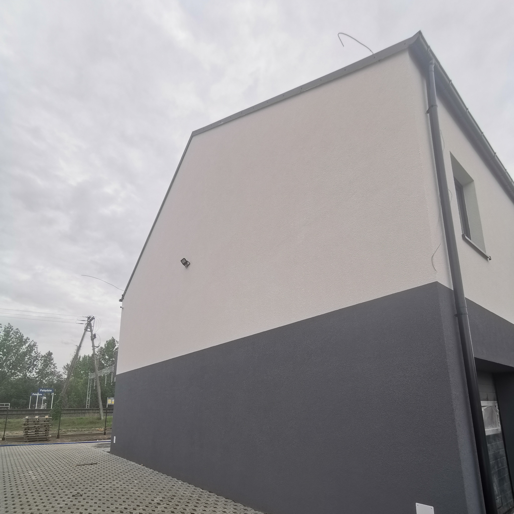 Nowoczesny dom z dwukolorową elewacją: górna część w jasnym odcieniu, dolna w ciemnoszarym, widoczny fragment okna, lampa elewacyjna i kostka brukowa przed budynkiem.