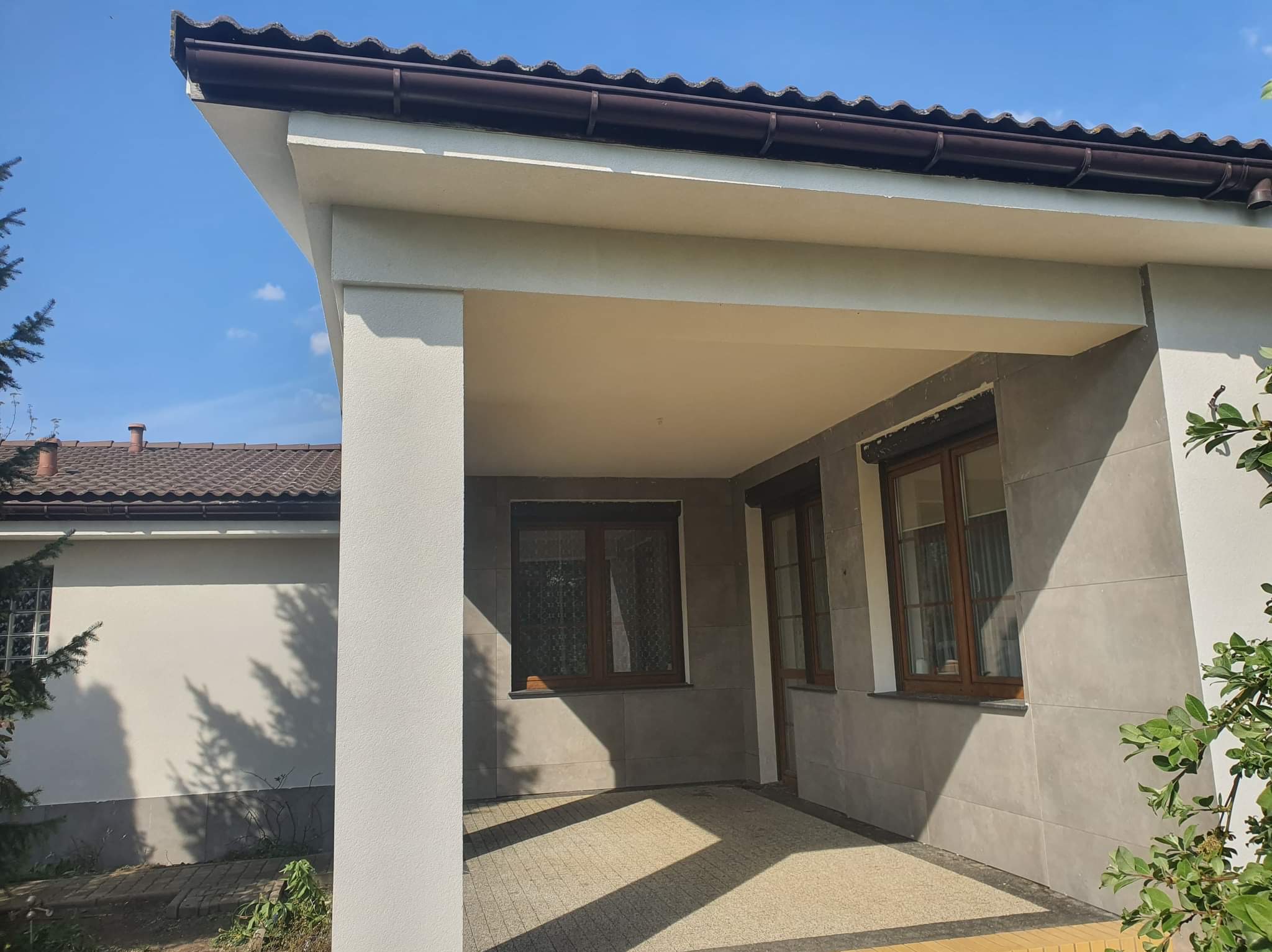 Zadaszone wejście do domu z szarą elewacją, brązowymi ramami okiennymi i żółtą kostką brukową na posadzce