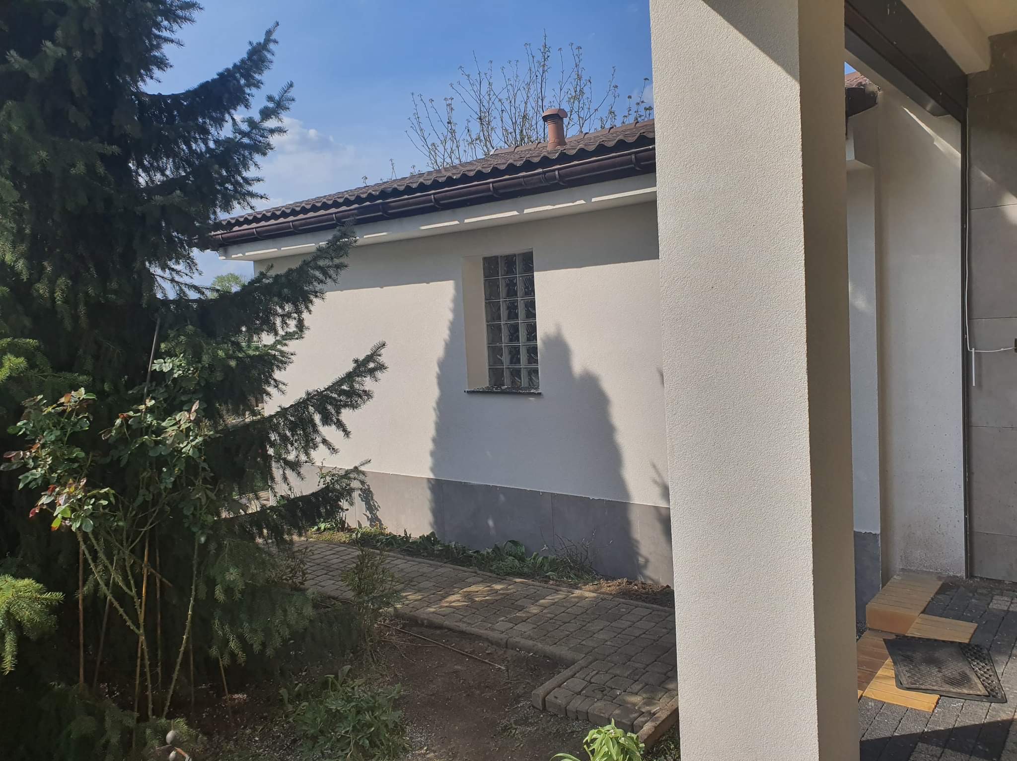 Boczny widok otynkowanej elewacji budynku z małym oknem z luksferów, z widoczną ścieżką z kostki brukowej i fragmentem iglastego drzewa, z uwzględnieniem cienia rzucanego przez drzewo.