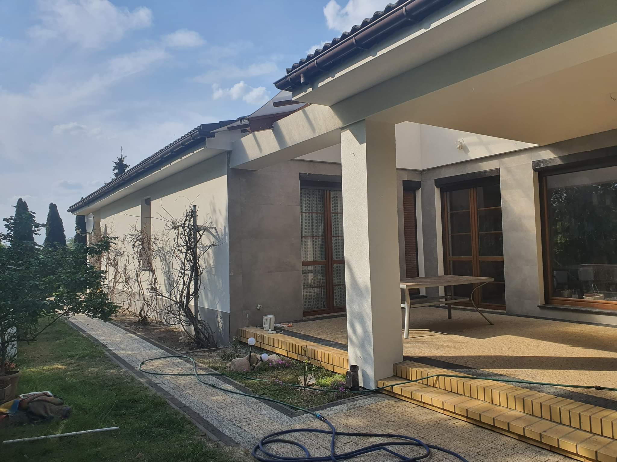 Jasna elewacja domu z zadaszonym tarasem, widoczne okna z drewnianymi ramami, kamienny chodnik i fragment ogrodu z roślinnością.