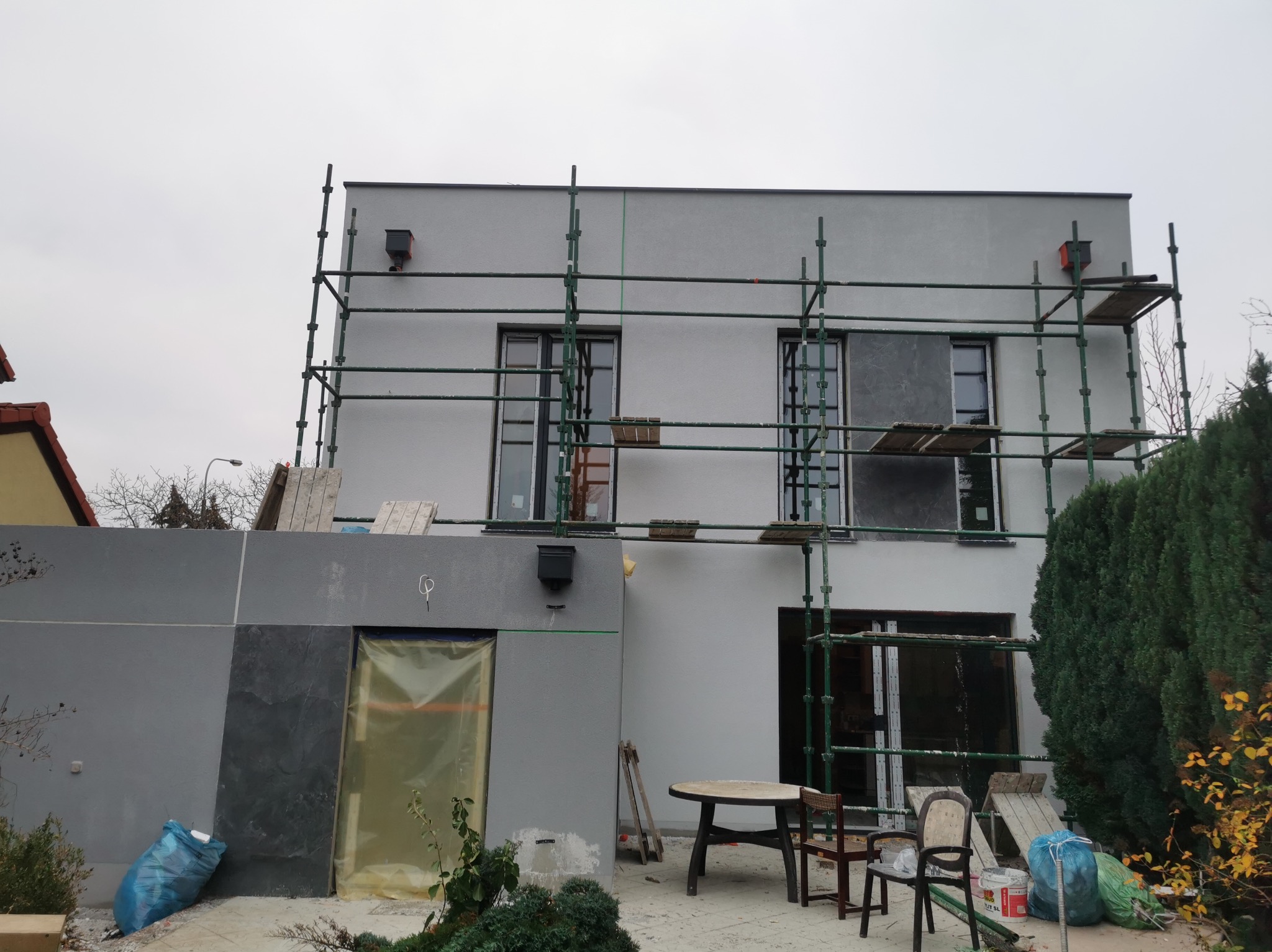 Dwukondygnacyjny dom w trakcie prac elewacyjnych, z ustawionymi rusztowaniami, częściowo otynkowany i pomalowany na szaro, z widocznymi zabezpieczeniami okien folią, elementami dekoracyjnymi...