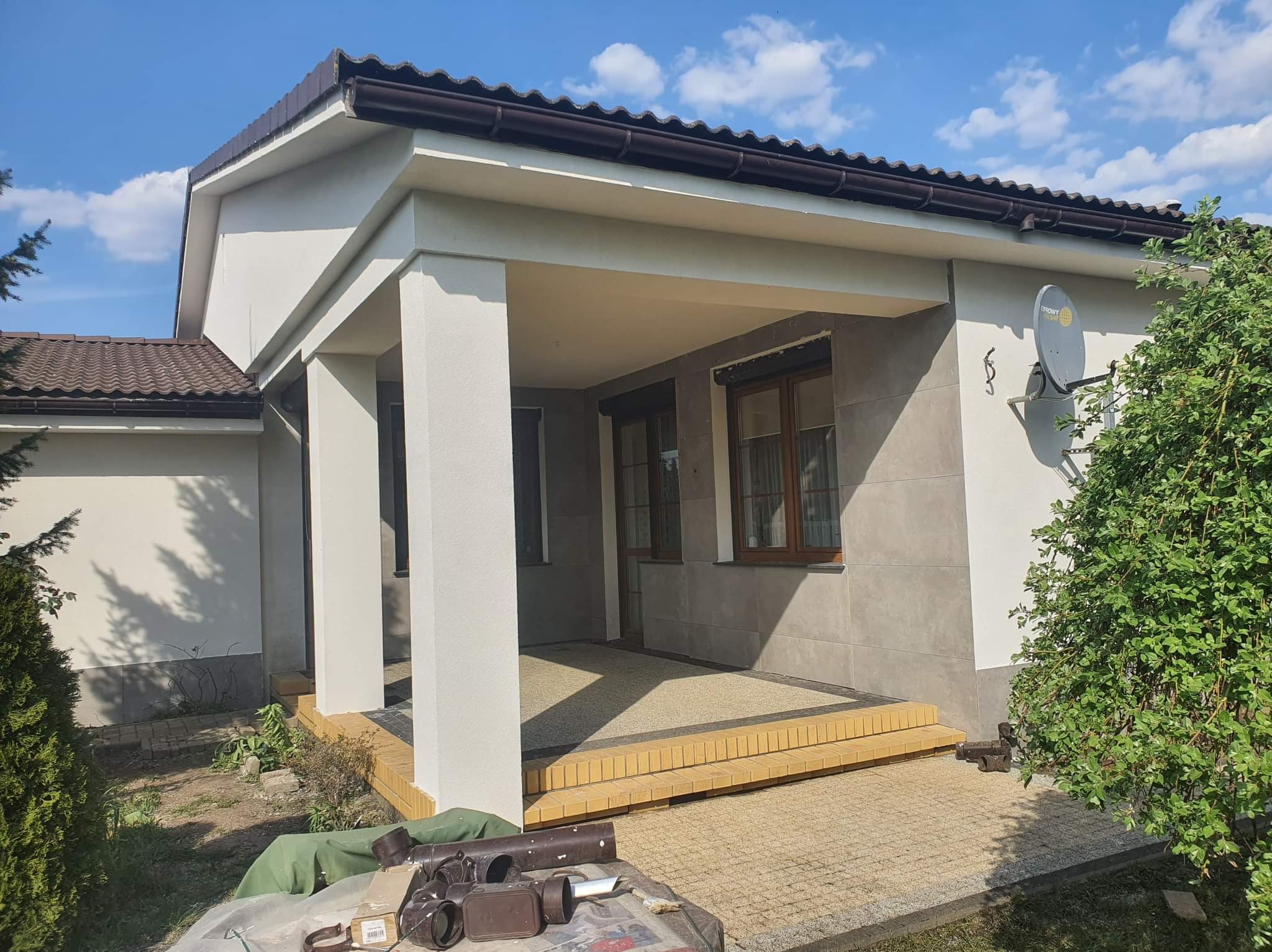 Wykończony taras z beżową kostką, schodami z żółtej cegły klinkierowej i szarą elewacją, widok na fragment domu z brązowym dachem i anteną satelitarną, w tle błękitne niebo.