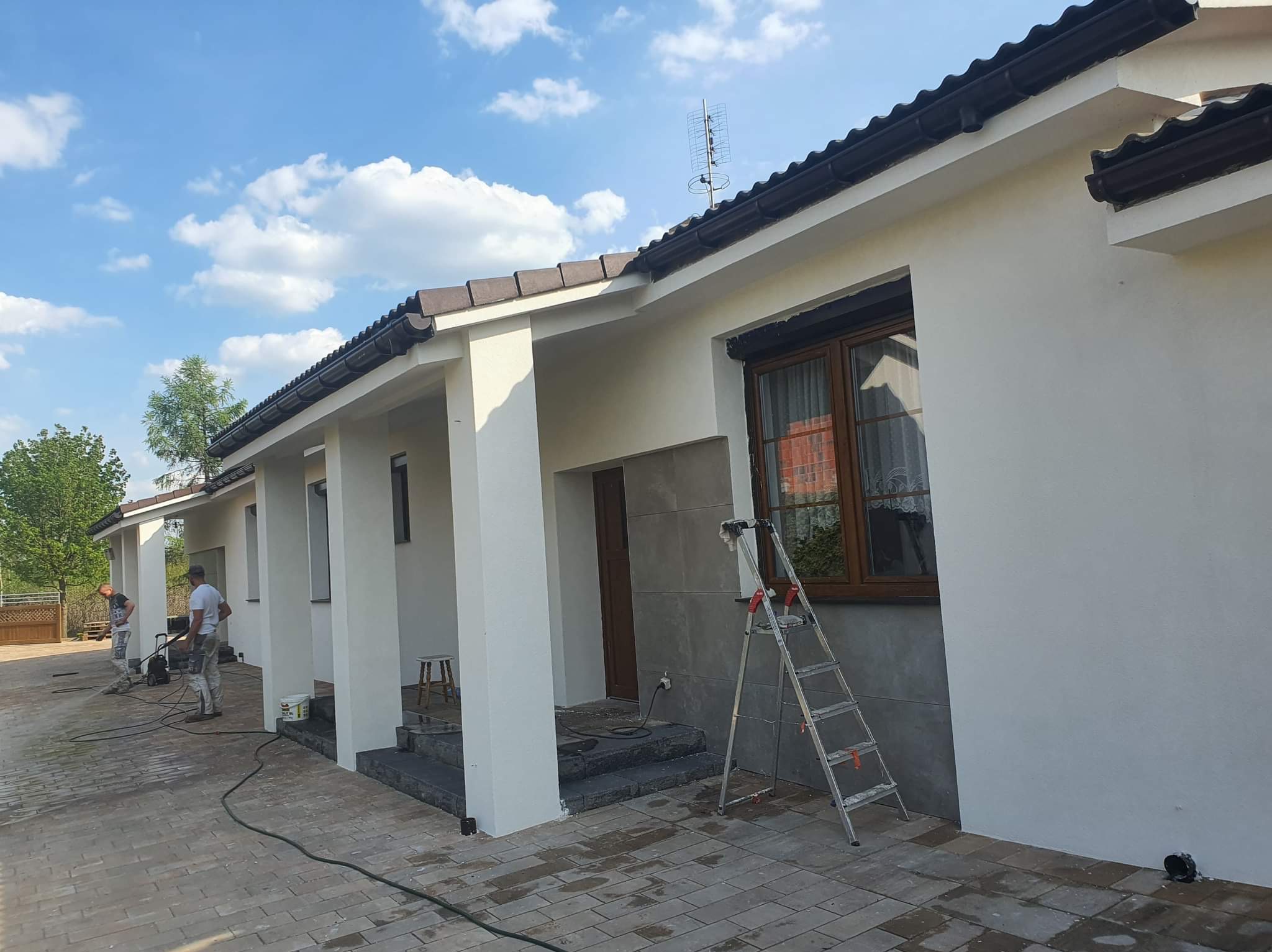 Dwóch mężczyzn maluje elewację domu z drabiny, widoczne narzędzia i sprzęt do malowania, błękitne niebo z chmurami w tle.