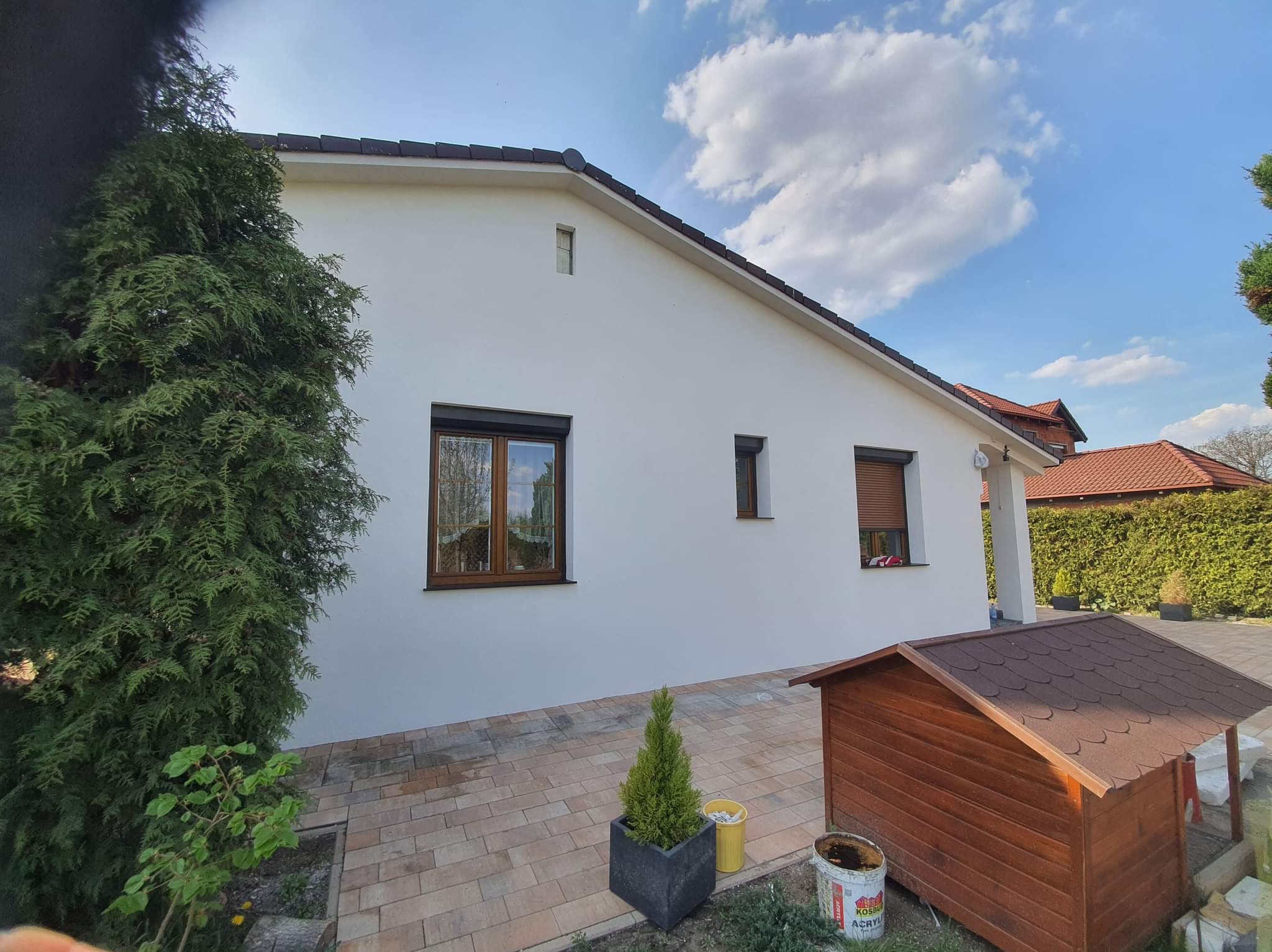 Biała elewacja domu jednorodzinnego po świeżym malowaniu, widoczne brązowe okna, brązowa dachówka, roślinność, kostka brukowa i wiaderko po farbie.