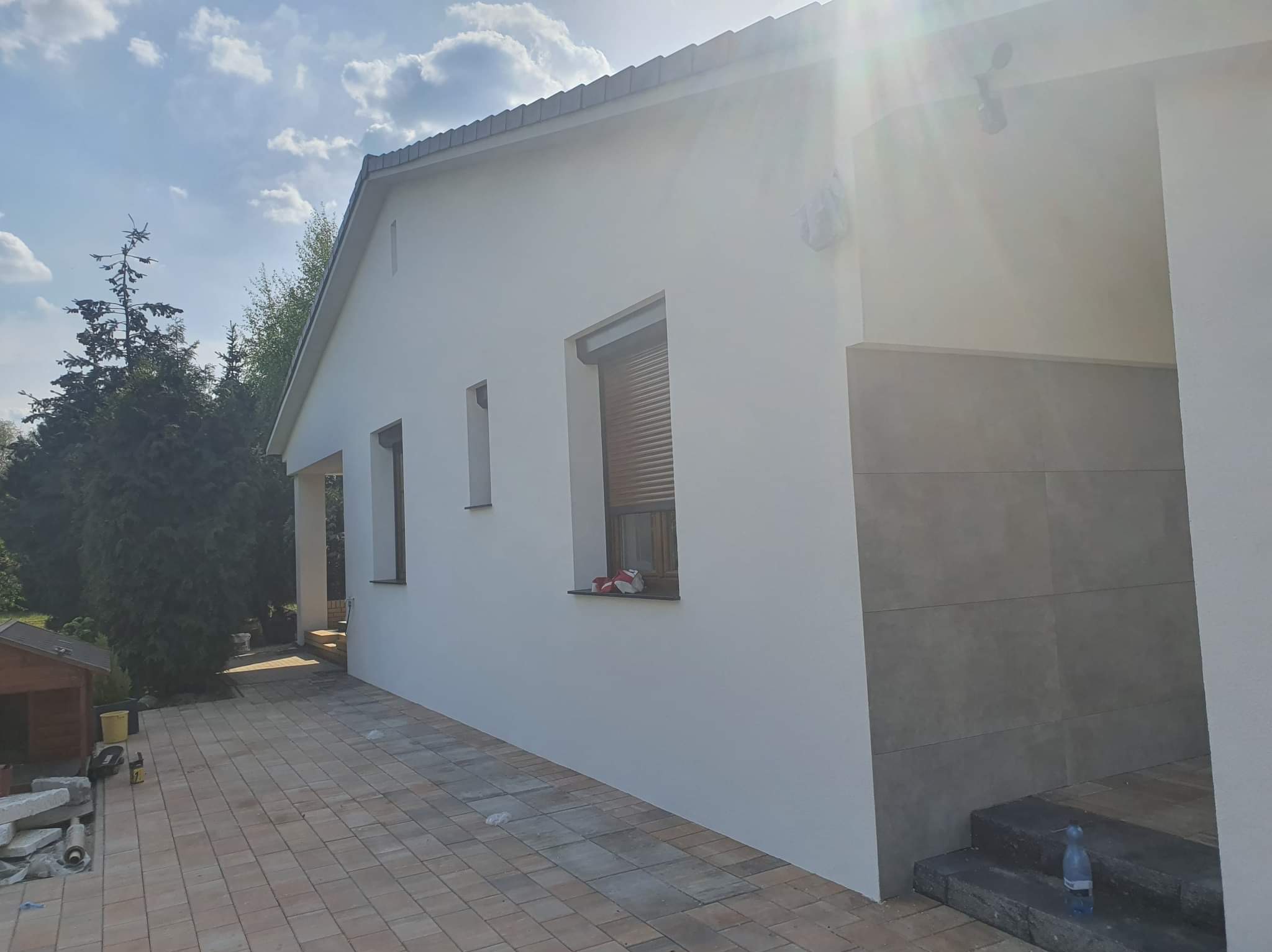 Biała elewacja domu jednorodzinnego po odświeżeniu, widoczne okna z częściowo zasłoniętymi roletami, oraz fragment tarasu z kostki brukowej. Na parapecie okna leżą czerwone rękawice robocze.