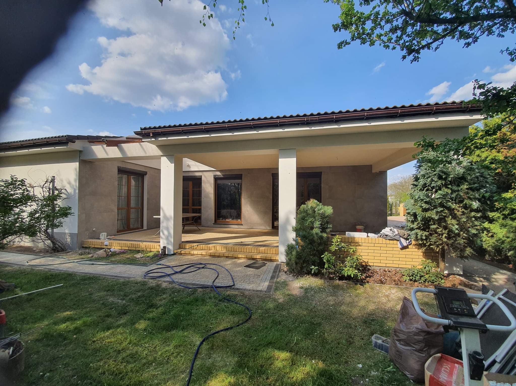 Niedokończona elewacja domu z odsłoniętym fragmentem ceglanej ściany, widoczne przewody i sprzęt budowlany na trawniku.