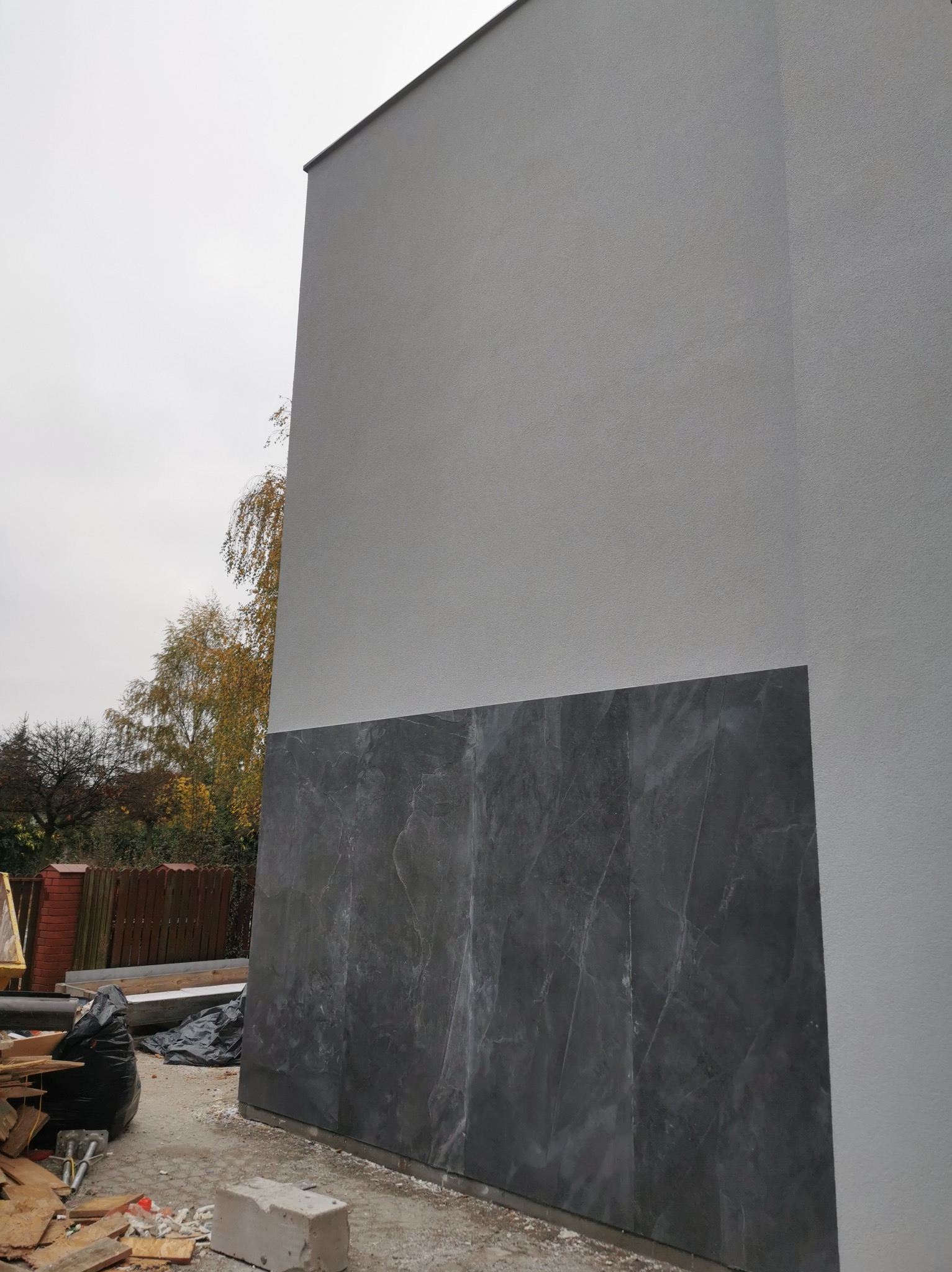 Fragment ściany budynku z częściowo położoną okładziną imitującą ciemny marmur, widoczne elementy budowlane i otoczenie w trakcie prac wykończeniowych.