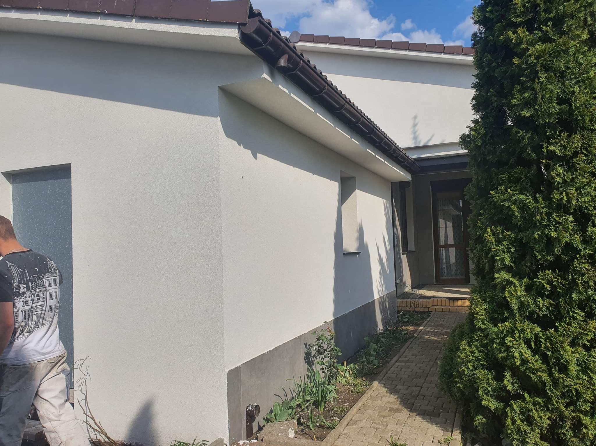 Świeżo pomalowana, jasna elewacja domu jednorodzinnego z brązowymi rynnami i fragmentem tui; widoczny pracownik w ubraniu roboczym przy oknie.