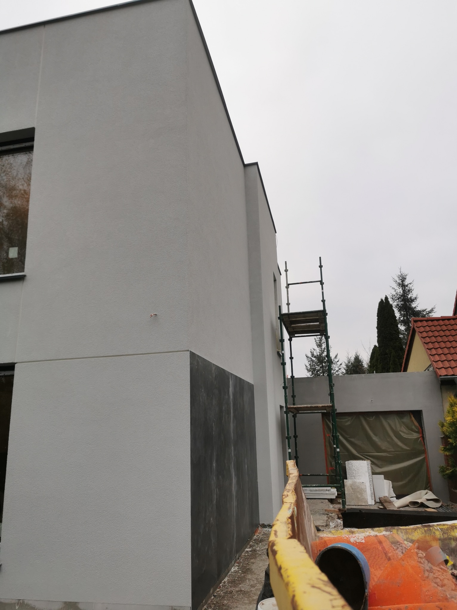 Nowoczesny dom z szarą elewacją w trakcie prac wykończeniowych, widoczne rusztowanie i materiały budowlane, fragment nieotynkowanej ściany.