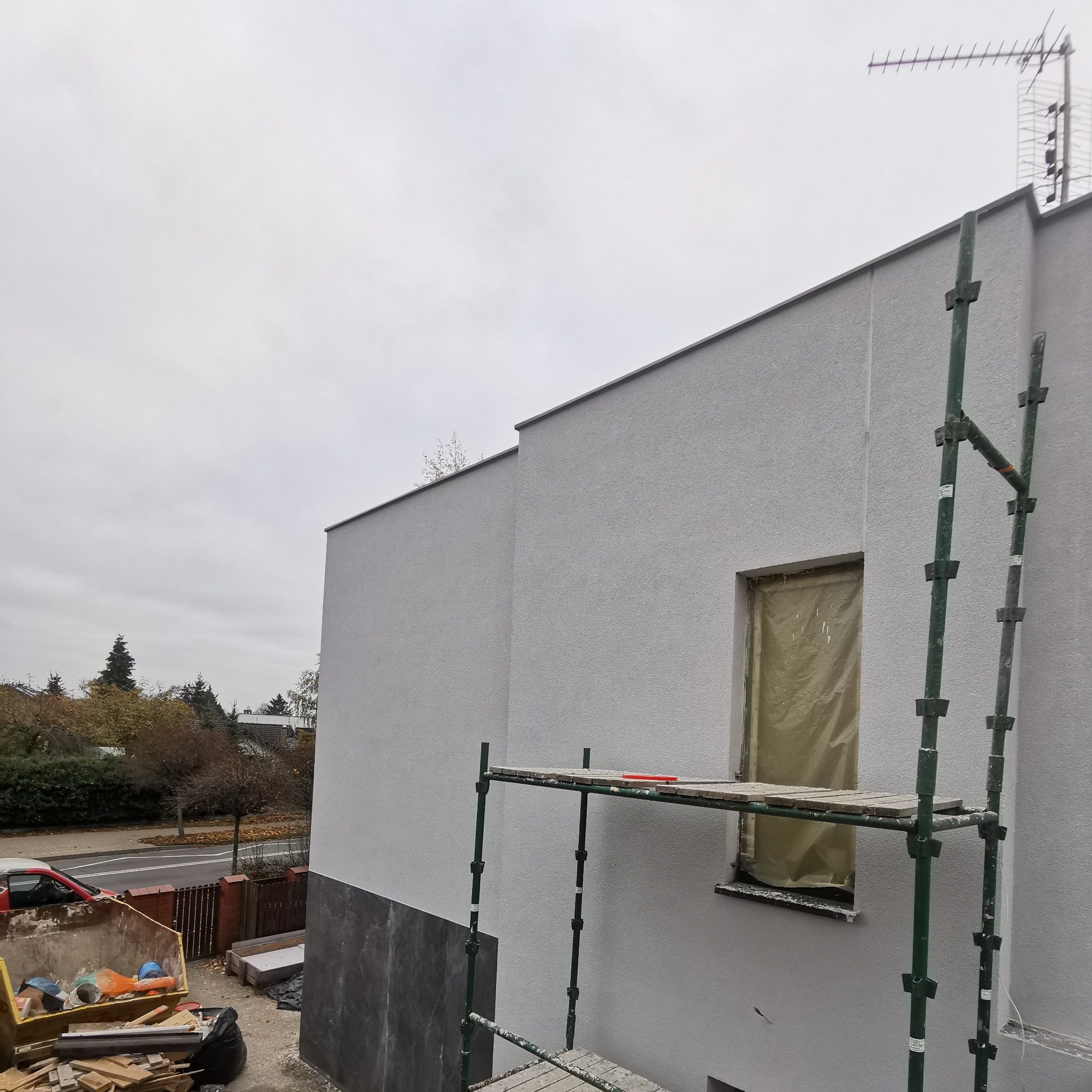 Świeżo otynkowana elewacja budynku z zabezpieczonym oknem, widoczne rusztowanie i kontener z odpadami budowlanymi.