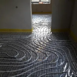 Białe rury ułożone w pętle na metalowej siatce, przygotowane do zalania betonem pod ogrzewanie podłogowe, widok z g&oacute;ry, ż&oacute;łta taśma wzdłuż ściany.