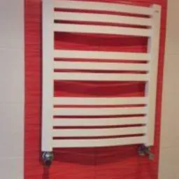 Biały, drabinkowy grzejnik łazienkowy zamontowany na czerwonej, błyszczącej ścianie z płytek, widoczne chromowane zawory.