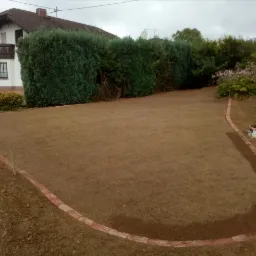 Przygotowany teren pod trawnik z obrzeżem z cegieł, widoczny dom w tle.